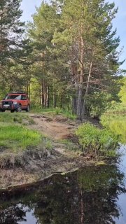 Красный внедорожник у берега в сосновом лесу Мещёры, видна песчаная тропа и тихая заводь с отражением деревьев на летнем пейзаже.