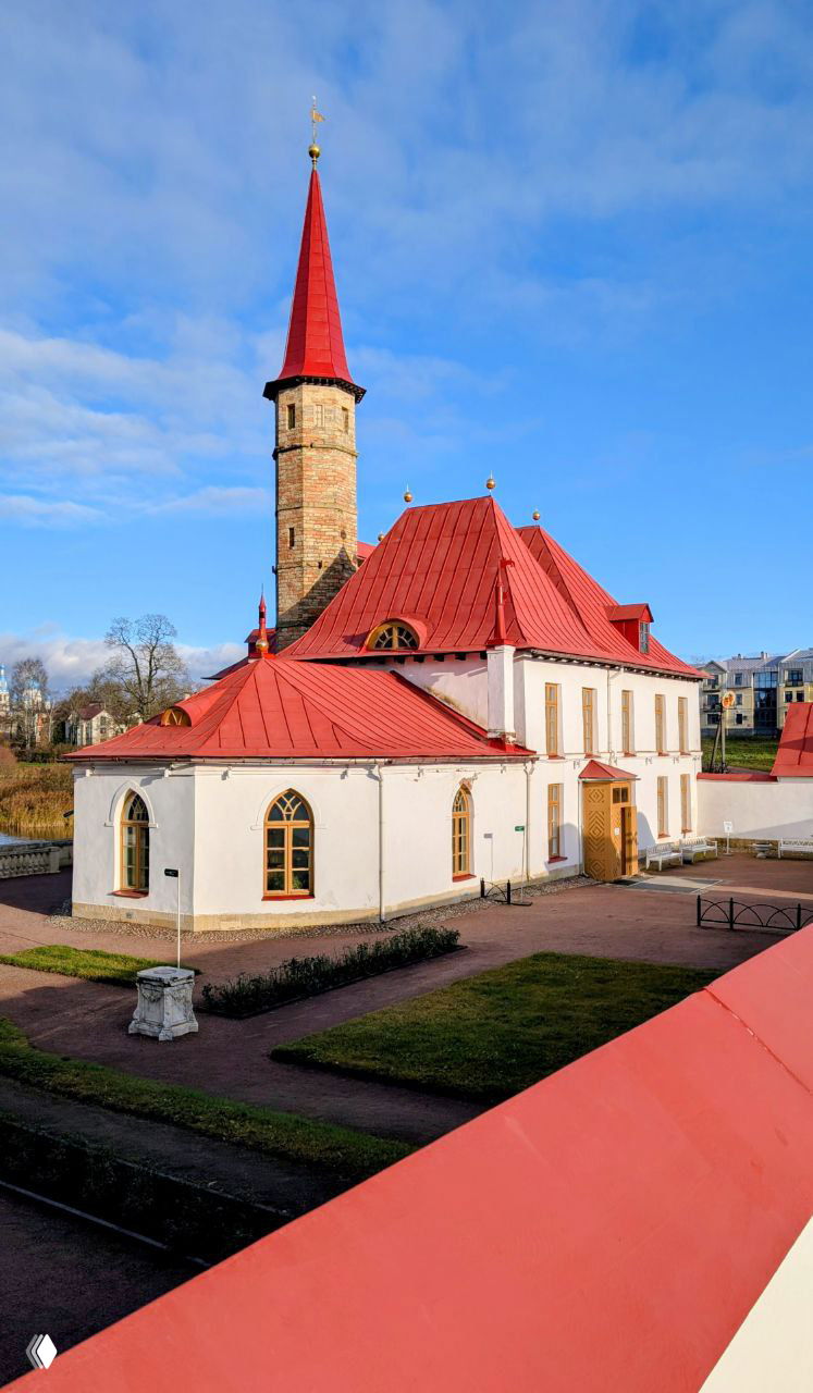 Фасад дворца с белыми стенами и красной острой крышей на фоне голубого неба; архитектурные готические элементы и аккуратный палисадник возле входа.