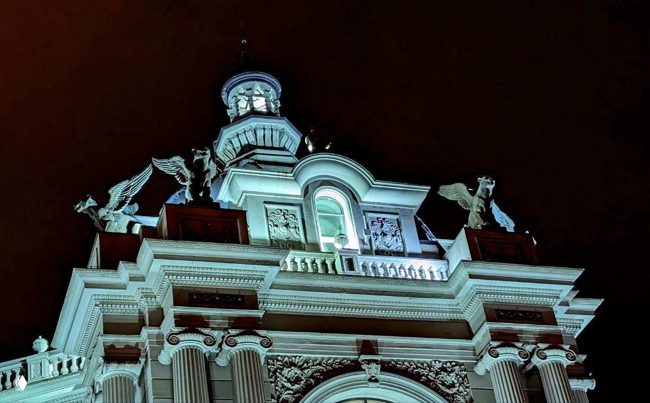 Крупный план верхней части фасада Дворца земледельцев с декоративными скульптурами и барельефами, подсветка подчёркивает рельеф.