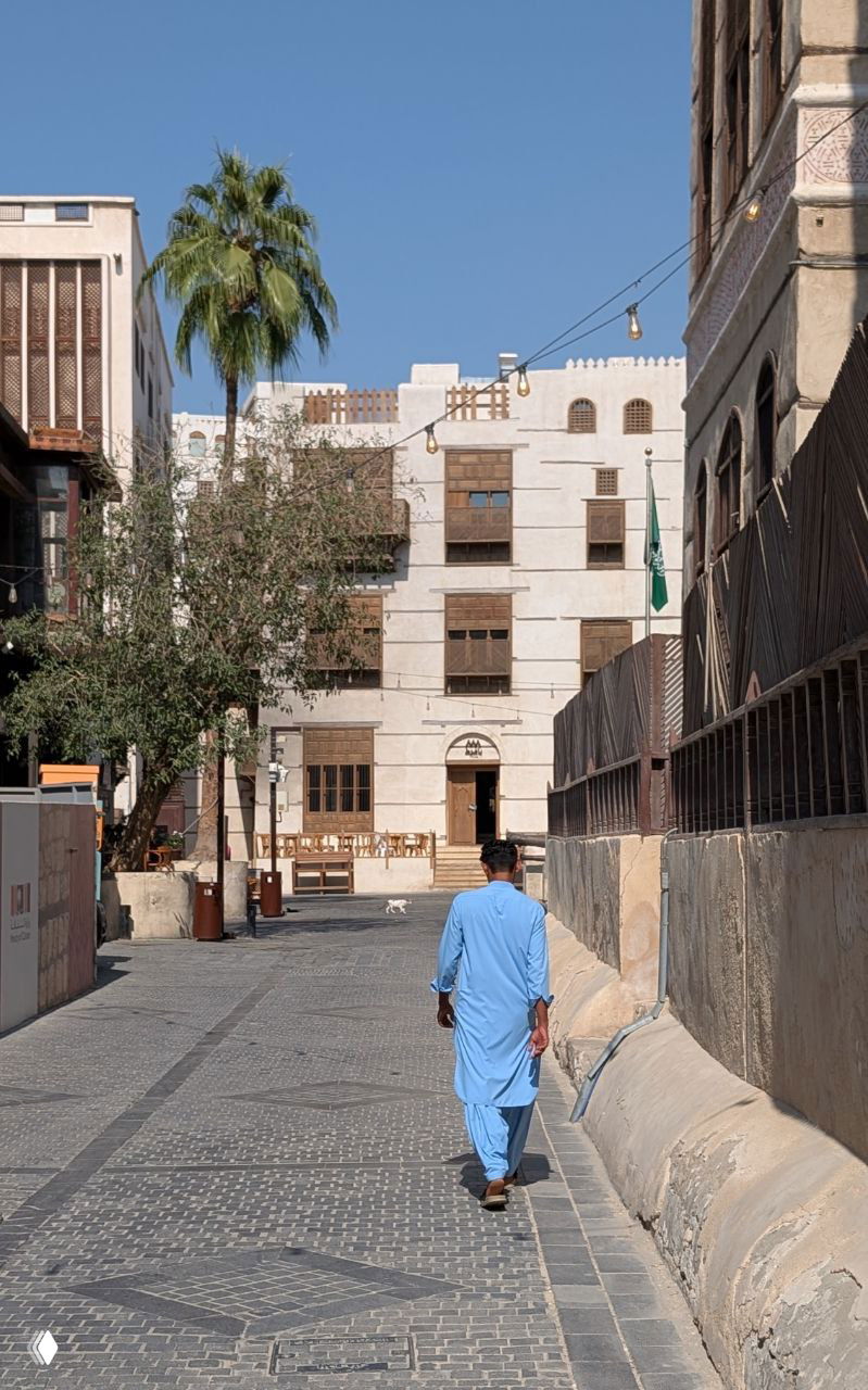 Пешеходная улица старой Джидды с узкими мощёными дорожками, один прохожий в традиционной одежде и здания с балконами на фоне.