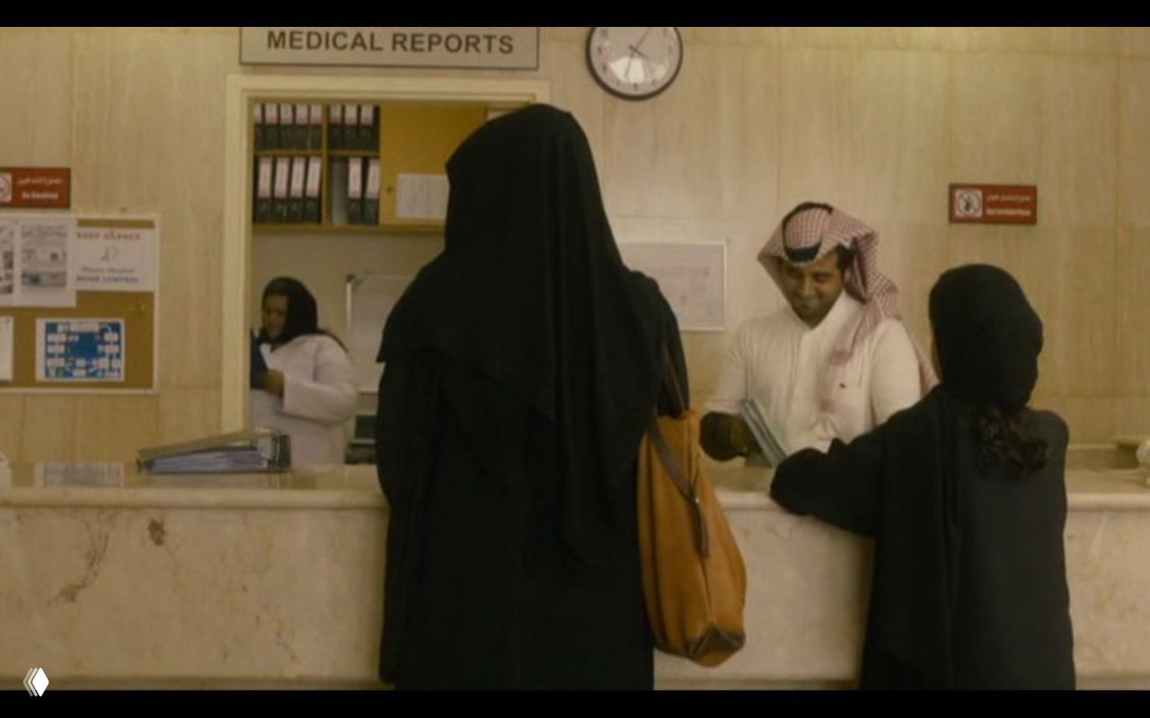Сцена в приёмной из «Ваджда»: женщины в абайях у стойки регистрации, мужчина-администратор — городской бытовой контекст и гендерные границы.