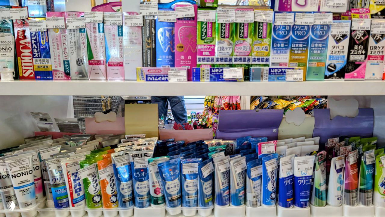 Полка магазина с множеством тюбиков зубной пасты и косметики, ценники и японский/корейский ассортимент товаров личной гигиены, общий план полки.