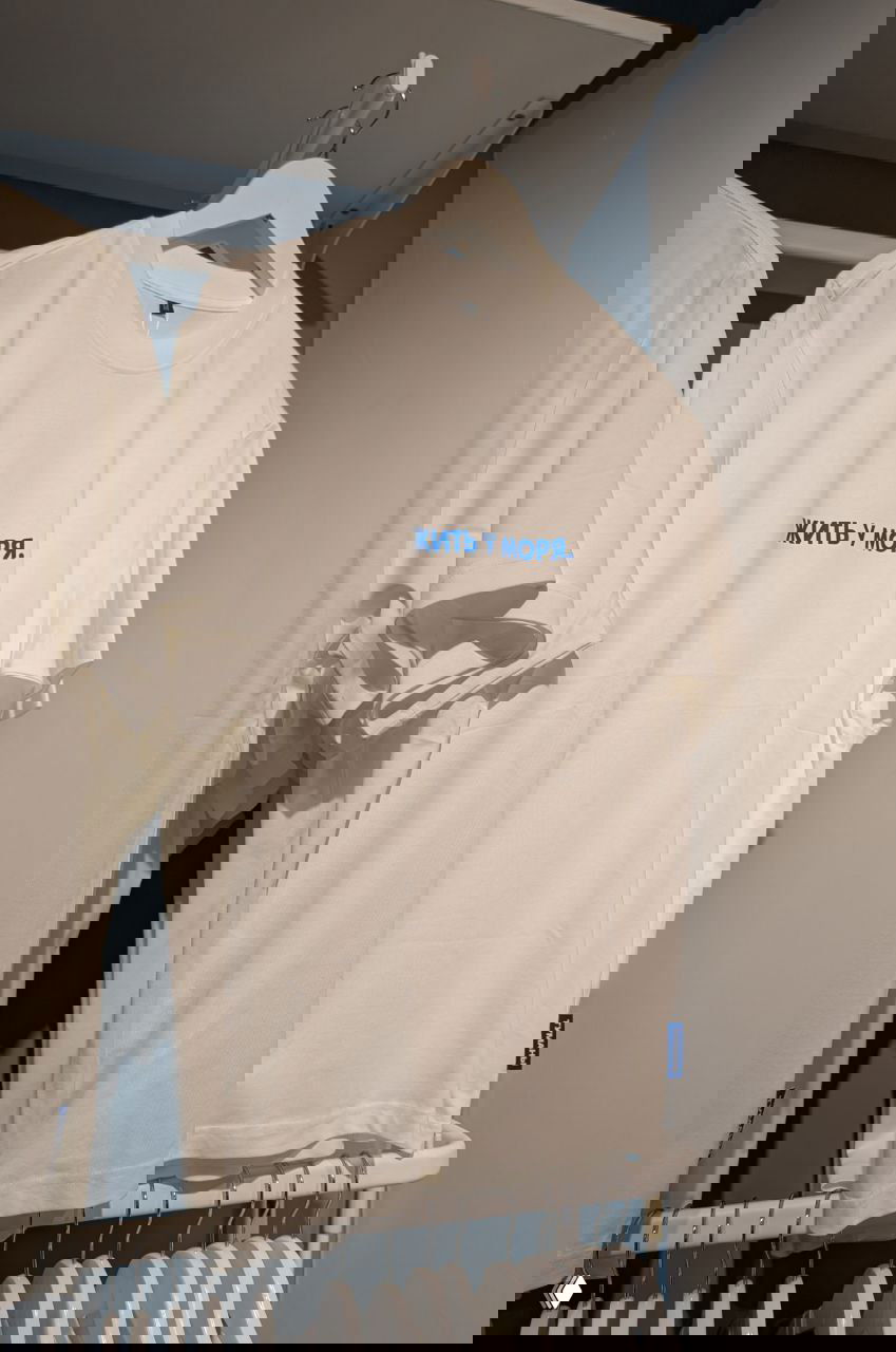 Белая футболка на вешалке с надписью синим шрифтом «ЖИТЬ У МОРЯ.», витрина или сувенирная лавка в аэропорту, предметная съёмка одежды.