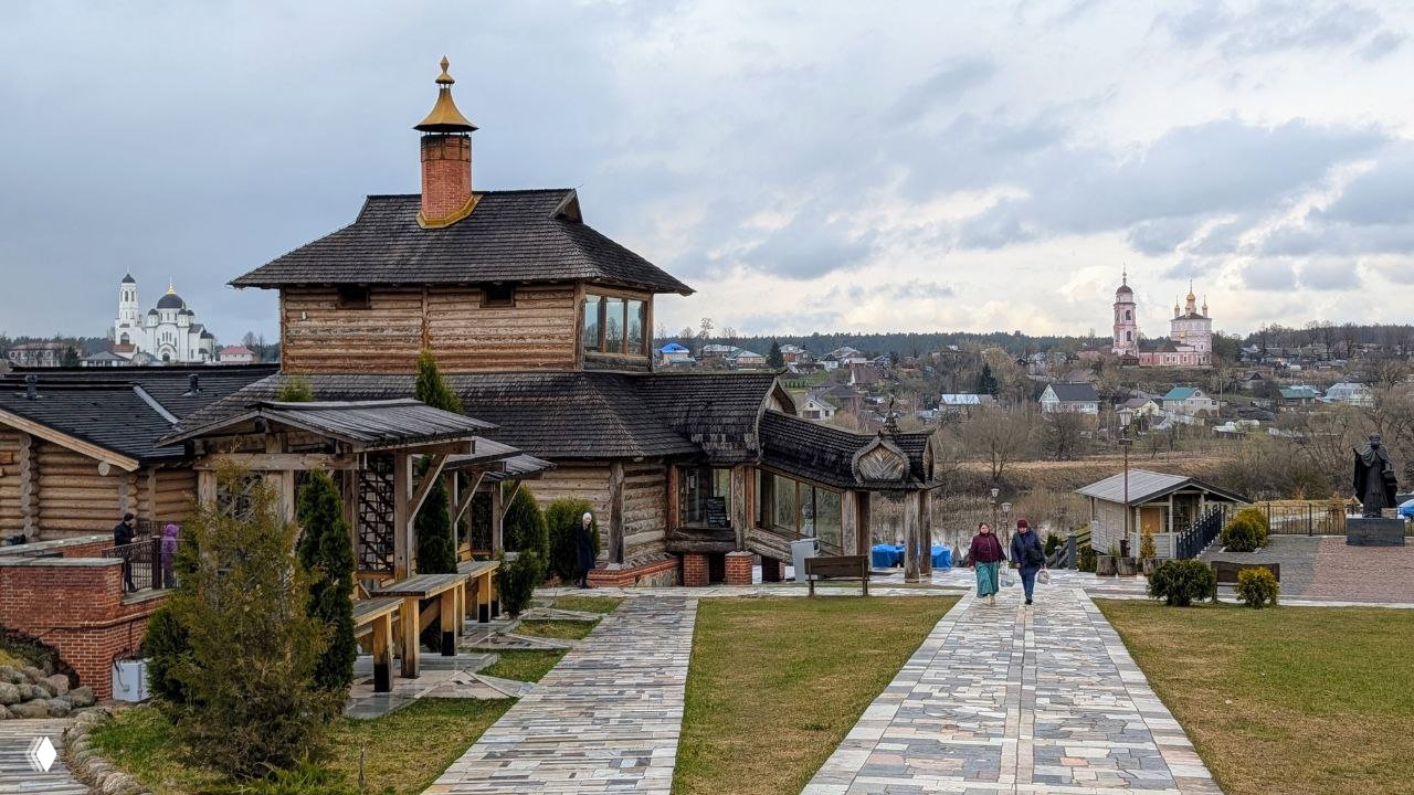 Территория у храма с деревянными постройками и аккуратно выложенной дорожкой, на горизонте видны купола и башни других храмов Боровска.