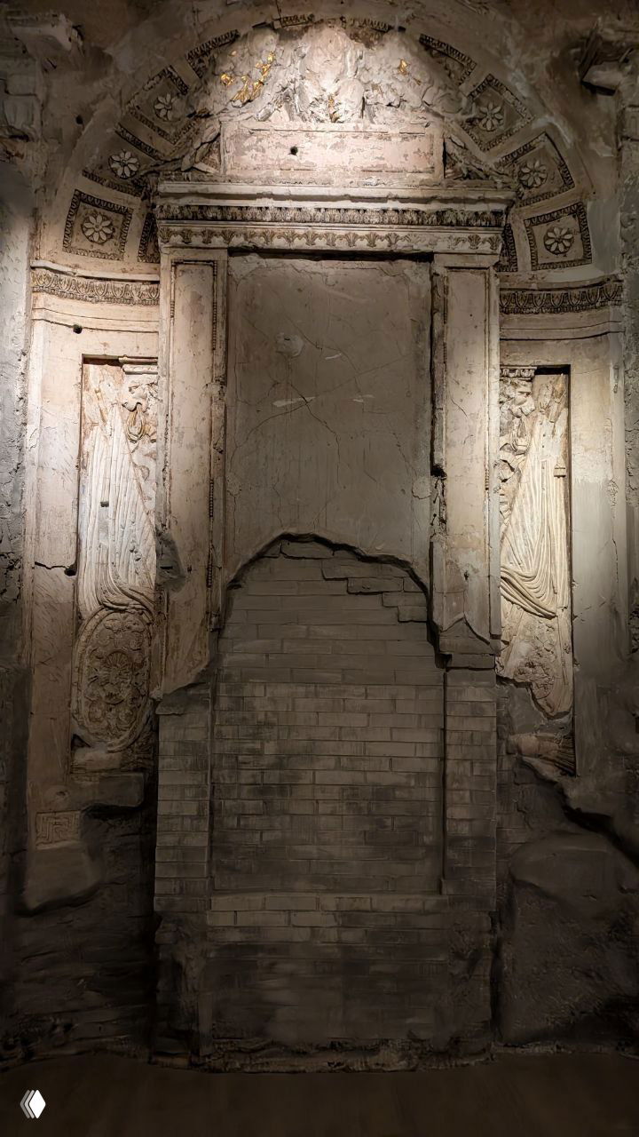 Интерьерный фрагмент: массивная дверь в светлом лаконичном стиле и побитая лепнина с барельефом на стене рядом, видны следы реставрации и расчищенные слои штукатурки.