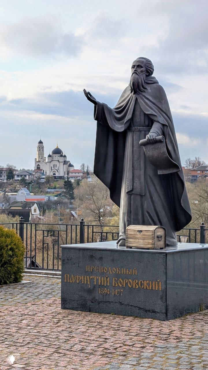 Памятник преподобному Пафнутию Борoвскому на постаменте, у ног которого деревянный сундучок, на заднем плане панорама Боровска и куполы собора.