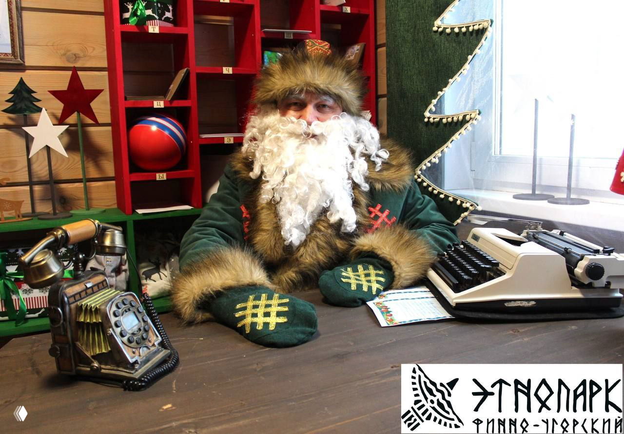 Резиденция местного Деда Мороза в Коми: мужчина в традиционном костюме с меховой шапкой и белой бородой сидит за столом в этнопарке.