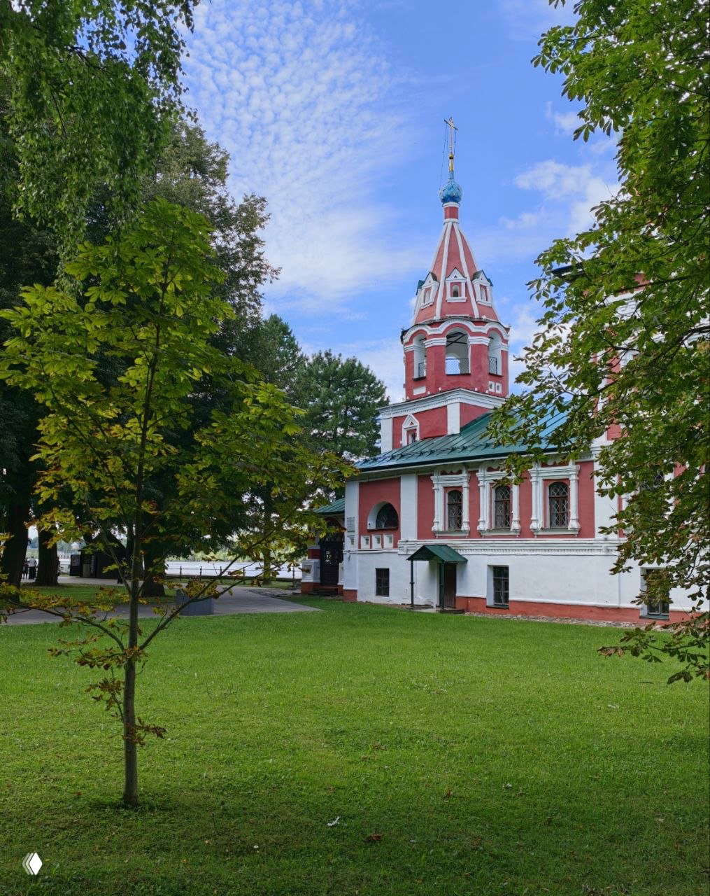 Красно-белая колокольня в парке Углича, окружённая лиственными деревьями и зелёным газоном на фоне голубого неба.