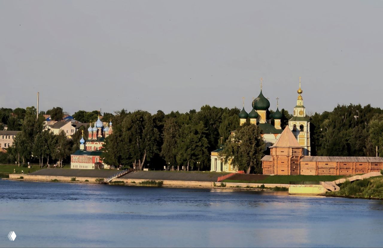 Вид через реку на набережную Углича: кремлёвские стены, купола собора и домики на фоне зелёных деревьев и спокойной воды.
