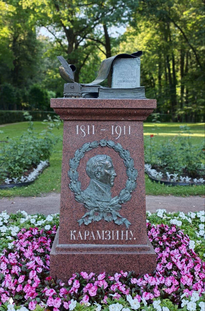 Памятник Карамзину на пьедестале с прорисованным профилем и датой 1811—1911, окружённый цветами на территории парка усадьбы Остафьево.
