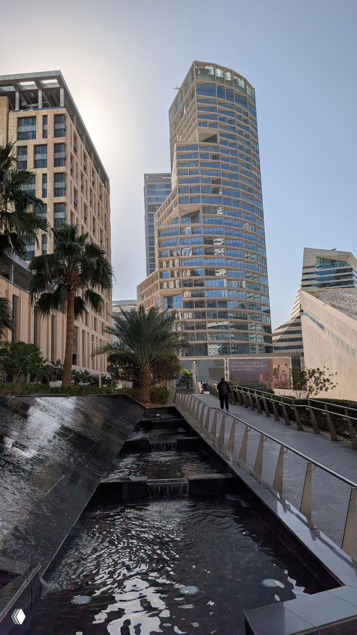 Современный городской пейзаж Эр-Рияда: высотное здание, пальмы и пешеходный мост с каскадной водной инсталляцией на переднем плане.