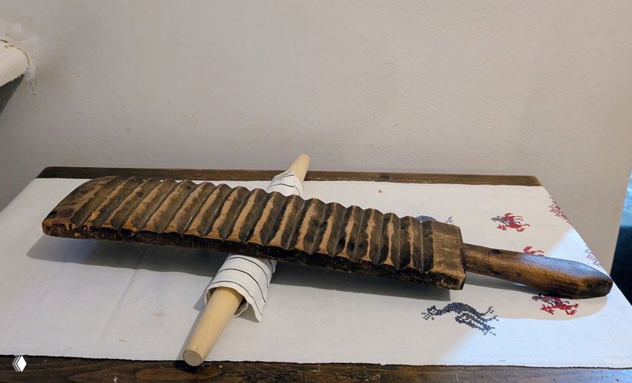 Деревянный рубель — старинный инструмент для глажки и маскировки, экспонат, лежащий на ткани с валиками в музейной витрине.