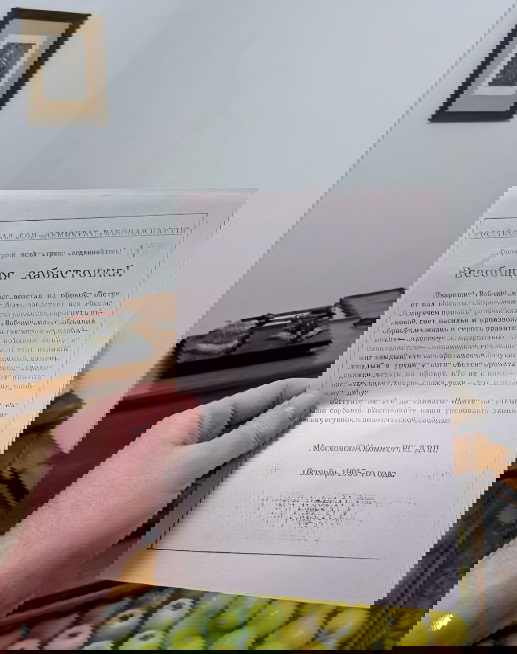 Рука держит репродукцию листовки 1905 года с заголовком «Всеобщая забастовка!», на заднем плане витрина с яблоками в музее.