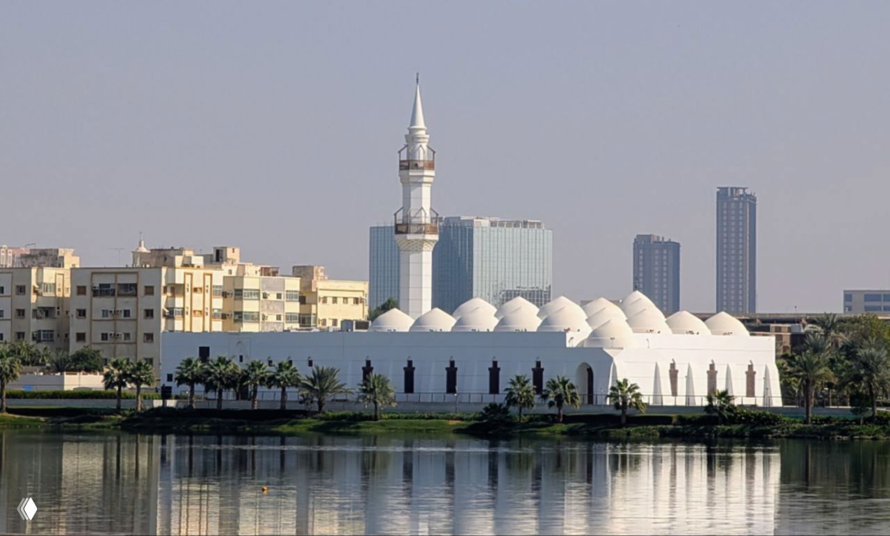 Белая мечеть у набережной: ряд куполов и минарет на фоне городских многоэтажек и спокойной воды, отражение в прибрежной лагуне.