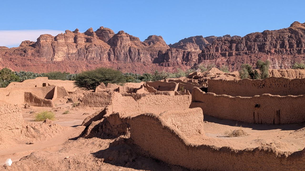 Пустынный пейзаж с красными скальными хребтами и остатками глинобитной деревни на переднем плане — северные горы и каньоны Саудовской Аравии, снято днём.