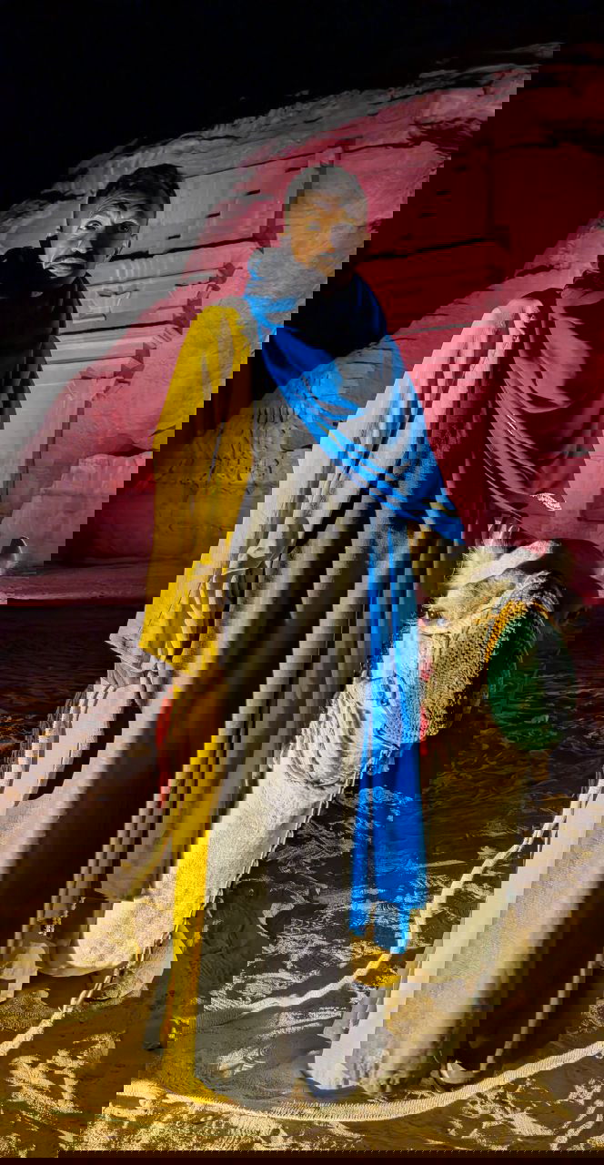 Человек в традиционной одежде рядом с верблюдом на фоне подсвеченных скал исторического комплекса — сценка, передающая атмосферу ночных шоу и туристических точек вроде АльУлы.