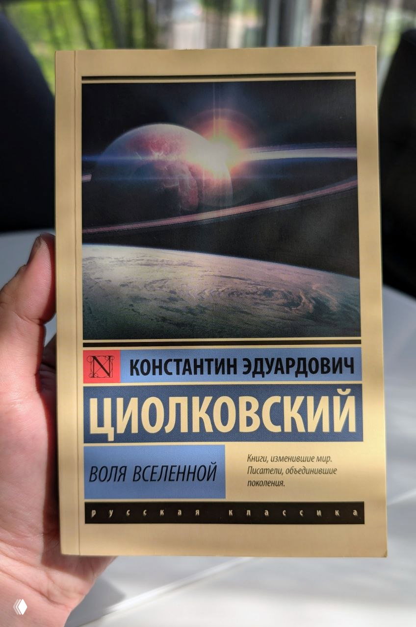 Рука держит книгу с космической обложкой: на титуле — «Константин Эдуардович Циолковский. Воля Вселенной», видна типографика.