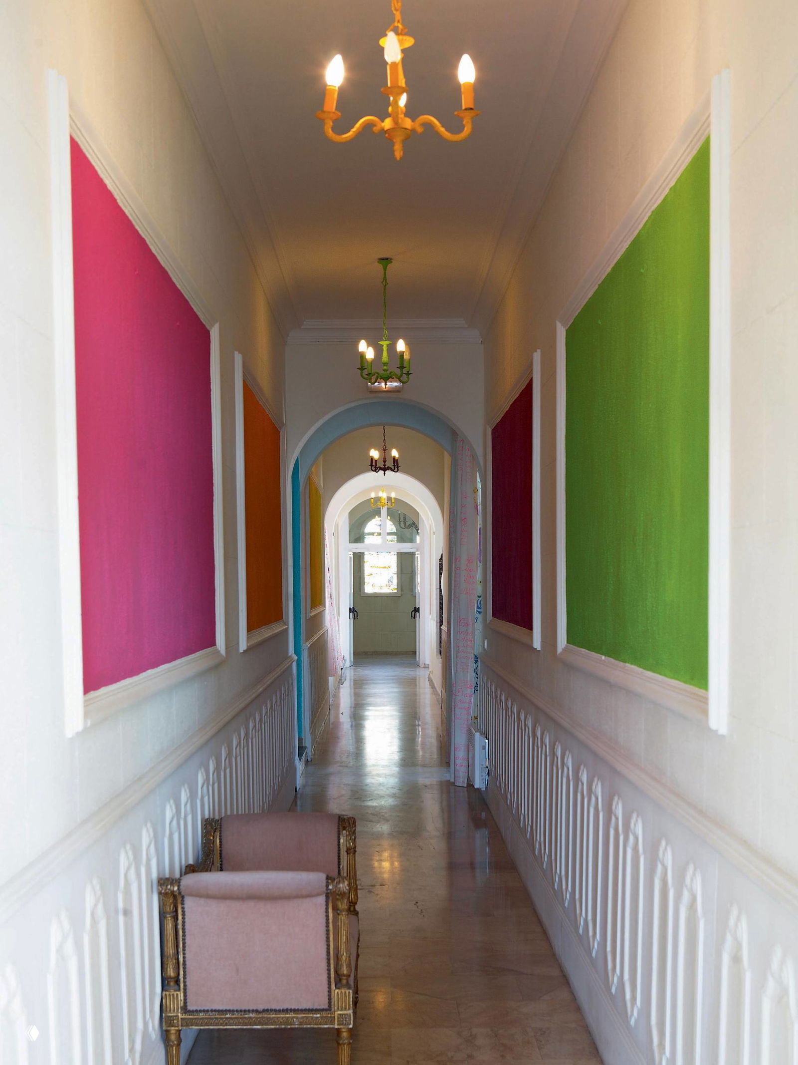 Длинный коридор с серией цветных панелей в рамках по бокам, арочные проходы и висящие люстры формируют эффектную перспективу пространства.