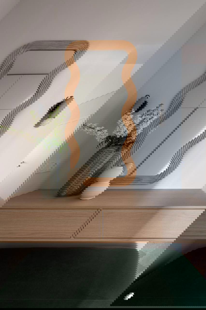 Прихожая: деревянная консоль с декоративными вазами, волнообразное зеркало в деревянной раме и темно-зеленая скамья под ней.