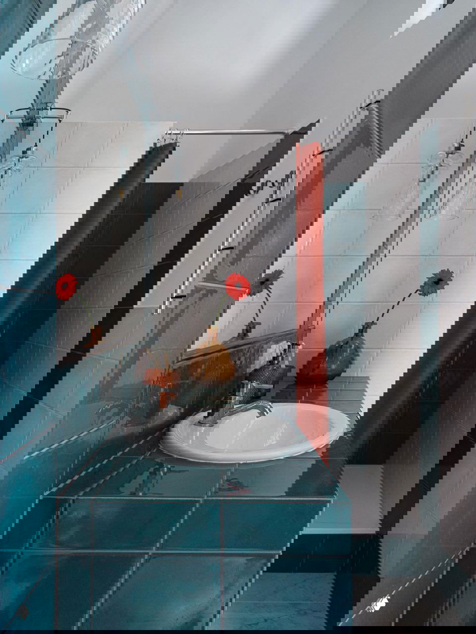 Ванная комната с глянцевой бирюзовой плиткой на стойке и стенах душевой, белая раковина и яркие цветочные акценты в виде красных цветов в вазе.