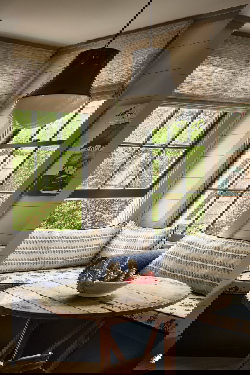 Уютная обеденная зона у окна с мягкими подушками на скамье, круглым деревянным столом и крупной подвесной лампой в тёплой палитре.