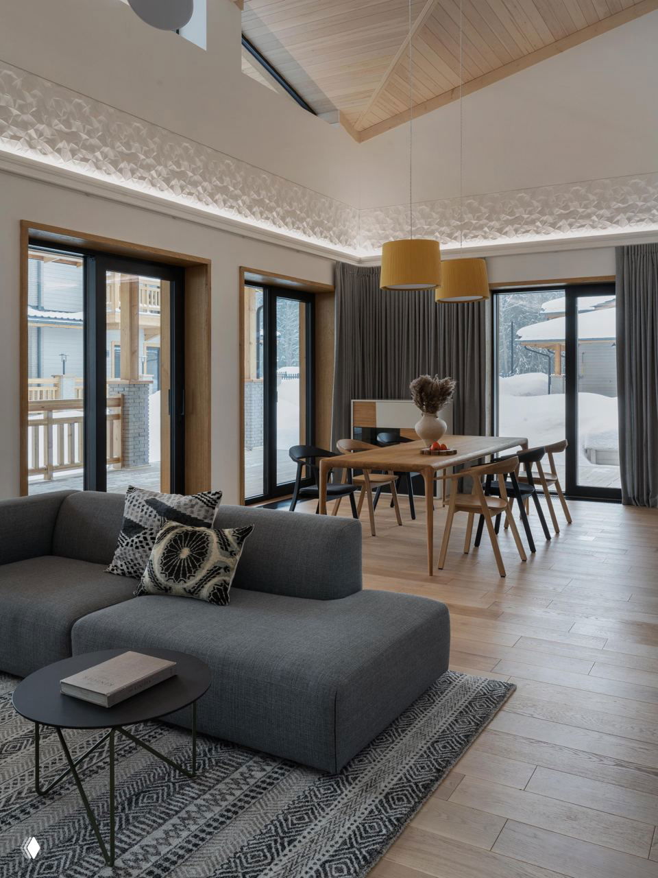 Просторная гостиная-столовая с высокими потолками: серый диван, деревянный стол, большие окна и нейтральный текстиль в спокойных тонах.