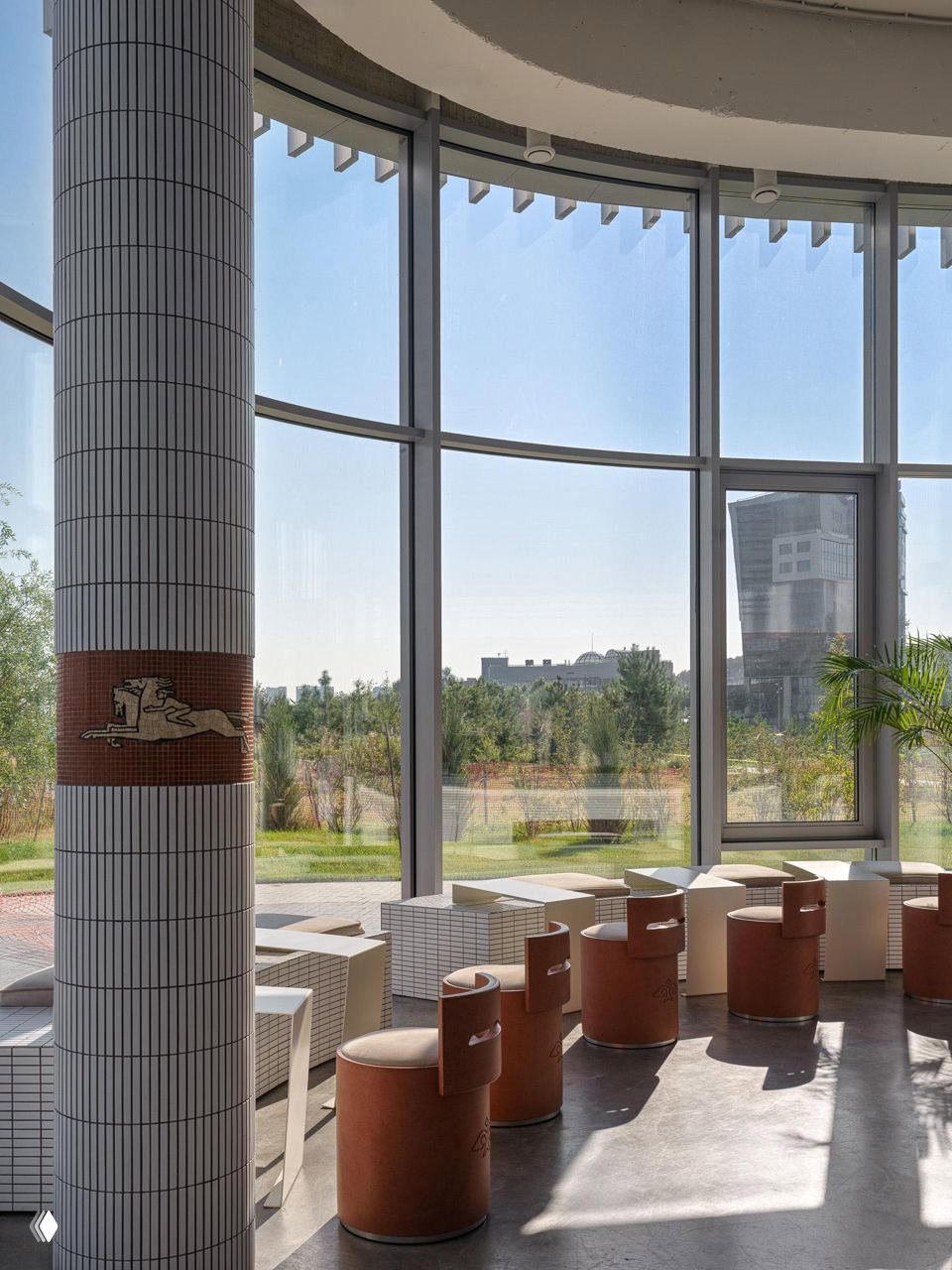 Зал с высокими окнами и колоннами, ряд круглых табуретов терракотового цвета и низкие столики на фоне городского ландшафта за стеклом.