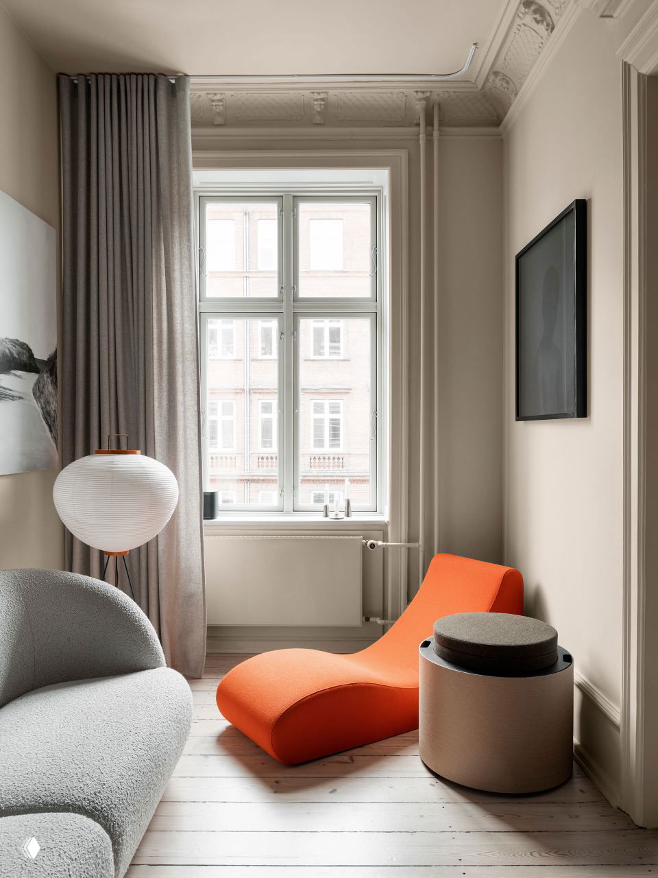 Маленькая комната с окном: яркая оранжевая шезлонга, светлый диван и круглый табурет на фоне нейтральных стен и светлого деревянного пола, спокойное дневное освещение.