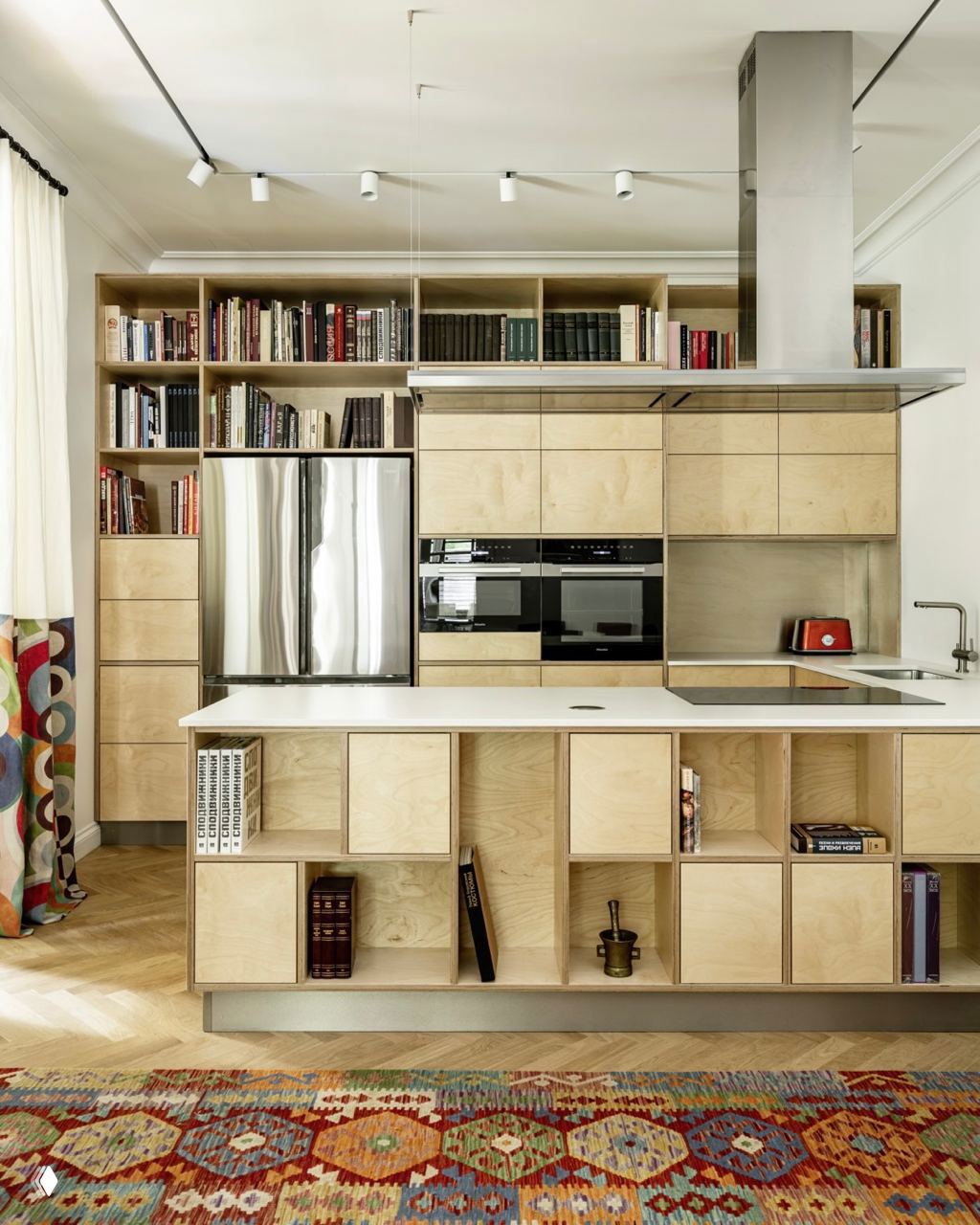 Кухонный гарнитур с фасадами из фанеры и встроенным холодильником; открытые полки над рабочей зоной и разноцветный ковёр создают тёплую эклектичную атмосферу интерьера.