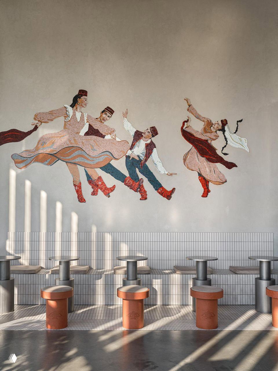 Стена с крупной мозаикой танцующих фигур в национальных костюмах над плиточной скамьёй и рядами круглых столиков в кафе.
