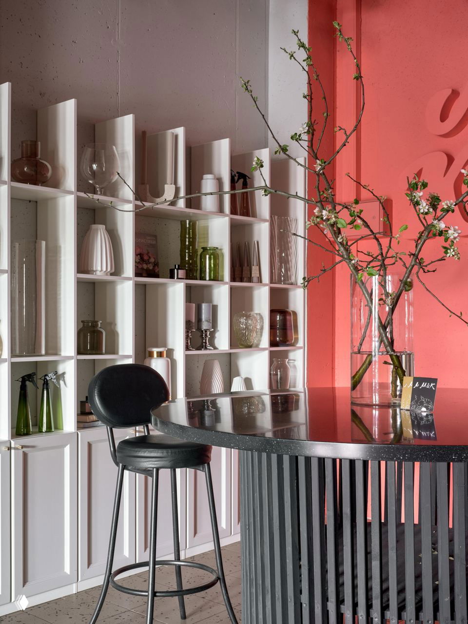 Крупный план стойки и полок с декоративными вазами и посудой, ветка цветущего растения на фоне коралловой стены.