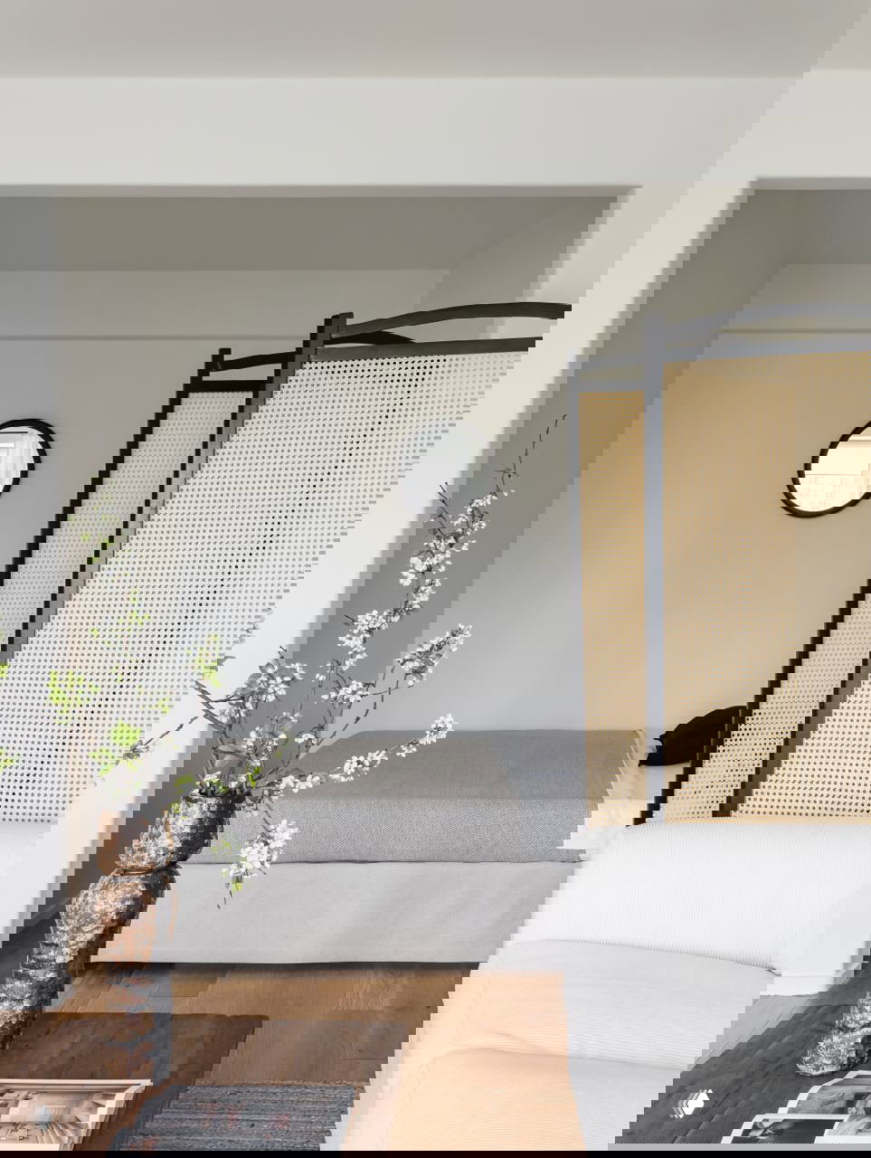 Ниша со спальным местом в студии: кровать за ширмой‑перегородкой из плетёной рейки, вид из гостиной, зелёные ветви в ваза на переднем плане.