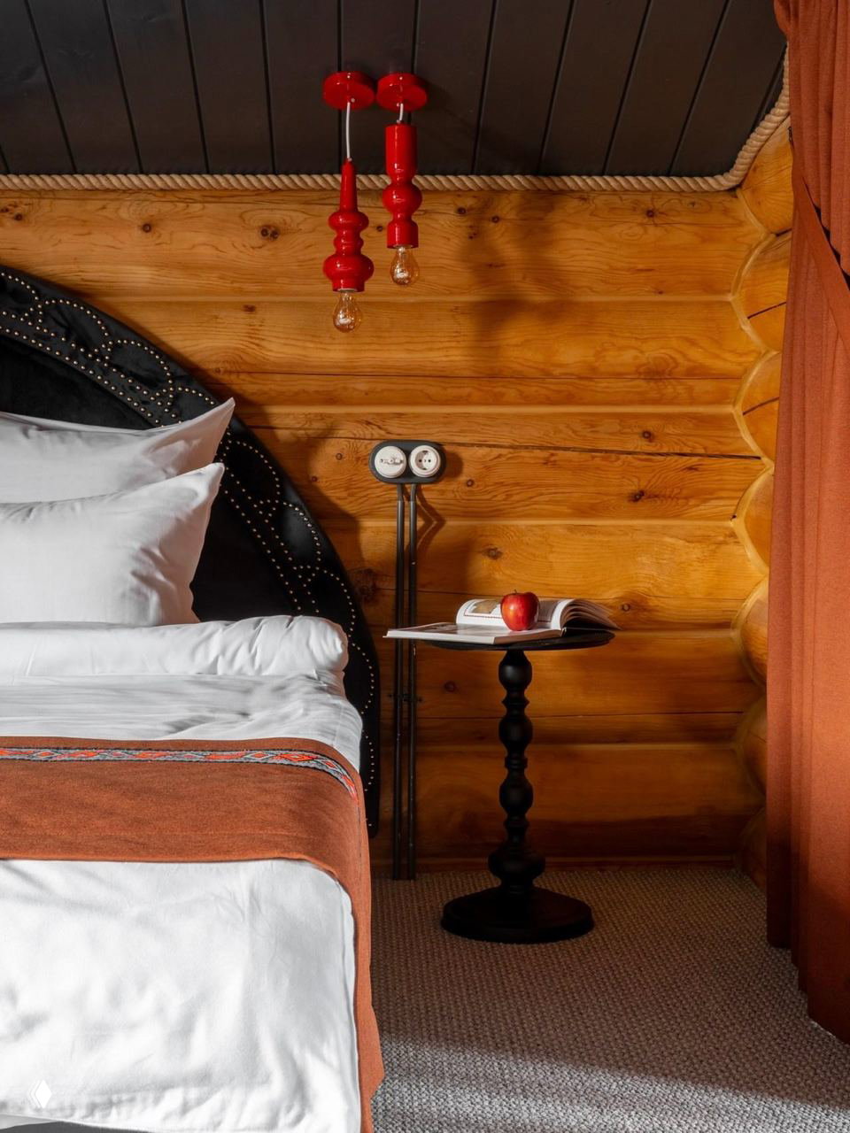 Крупный план спальни: резная деревянная стена из кедра, кровать с белым постельным бельём и красными текстильными акцентами, настенный бра и журнальный столик