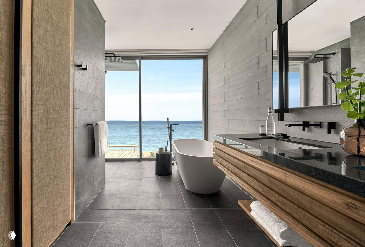 Ванная комната с видом на океан: ванна, тёмная плитка и большие стеклянные окна, выходящие к террасе и морю.