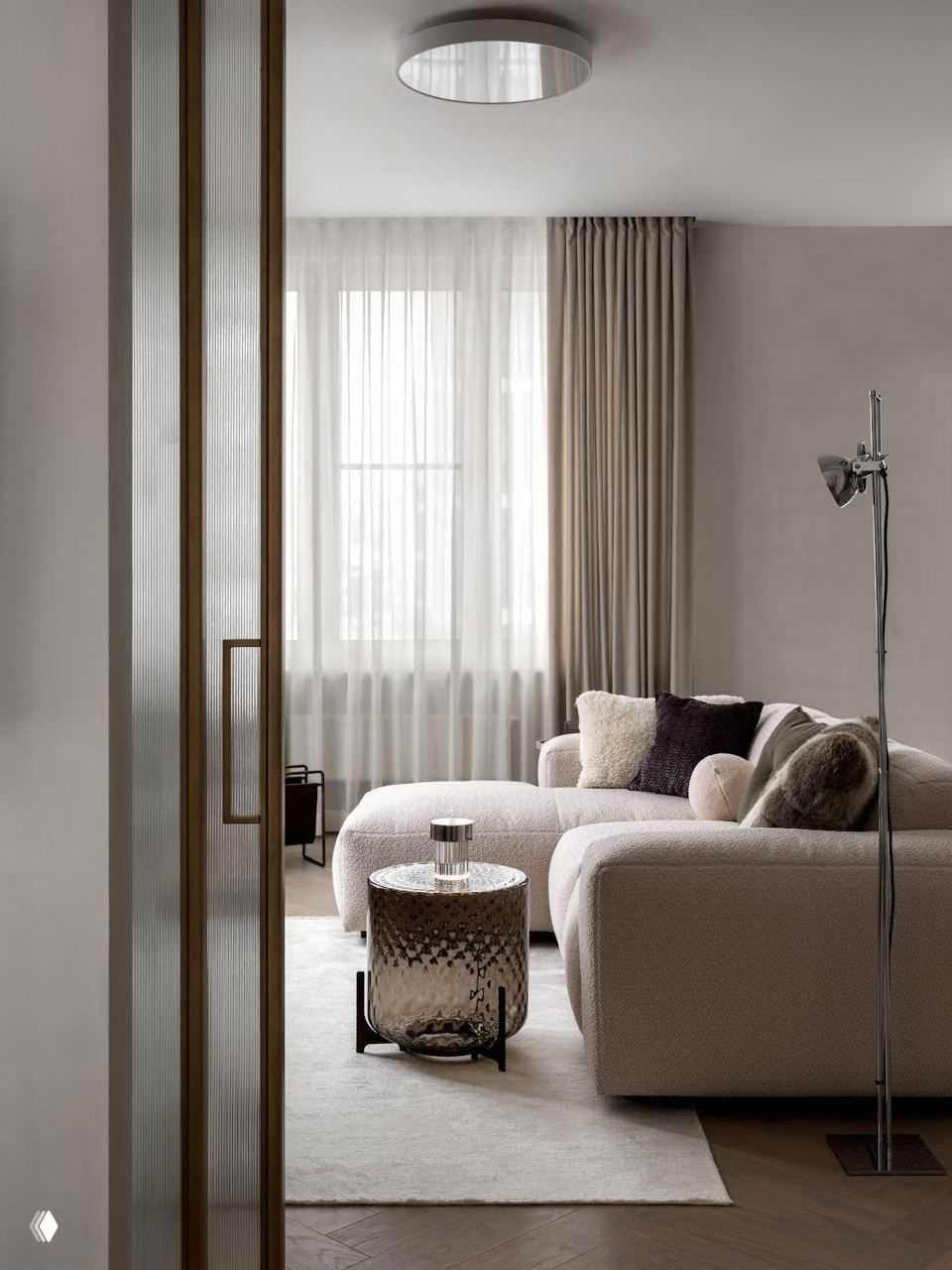 Гостиная через стеклянную перегородку: мягкий бежевый диван, подушки и тонкие шторы на окне — уютный минимализм в пастельной палитре проекта.
