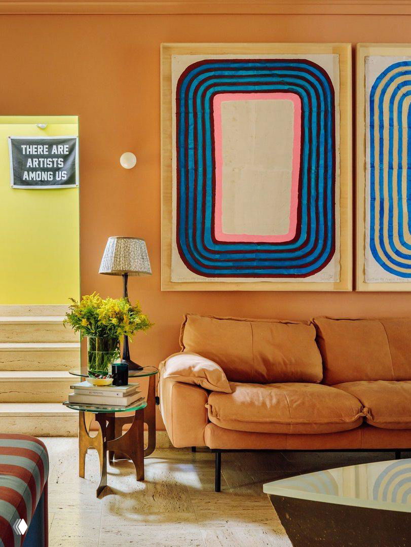 Гостиная с терракотовой стеной и оранжевым диваном, рядом крупные абстрактные картины и лимонно‑жёлтый проём — насыщенные цветовые акценты.