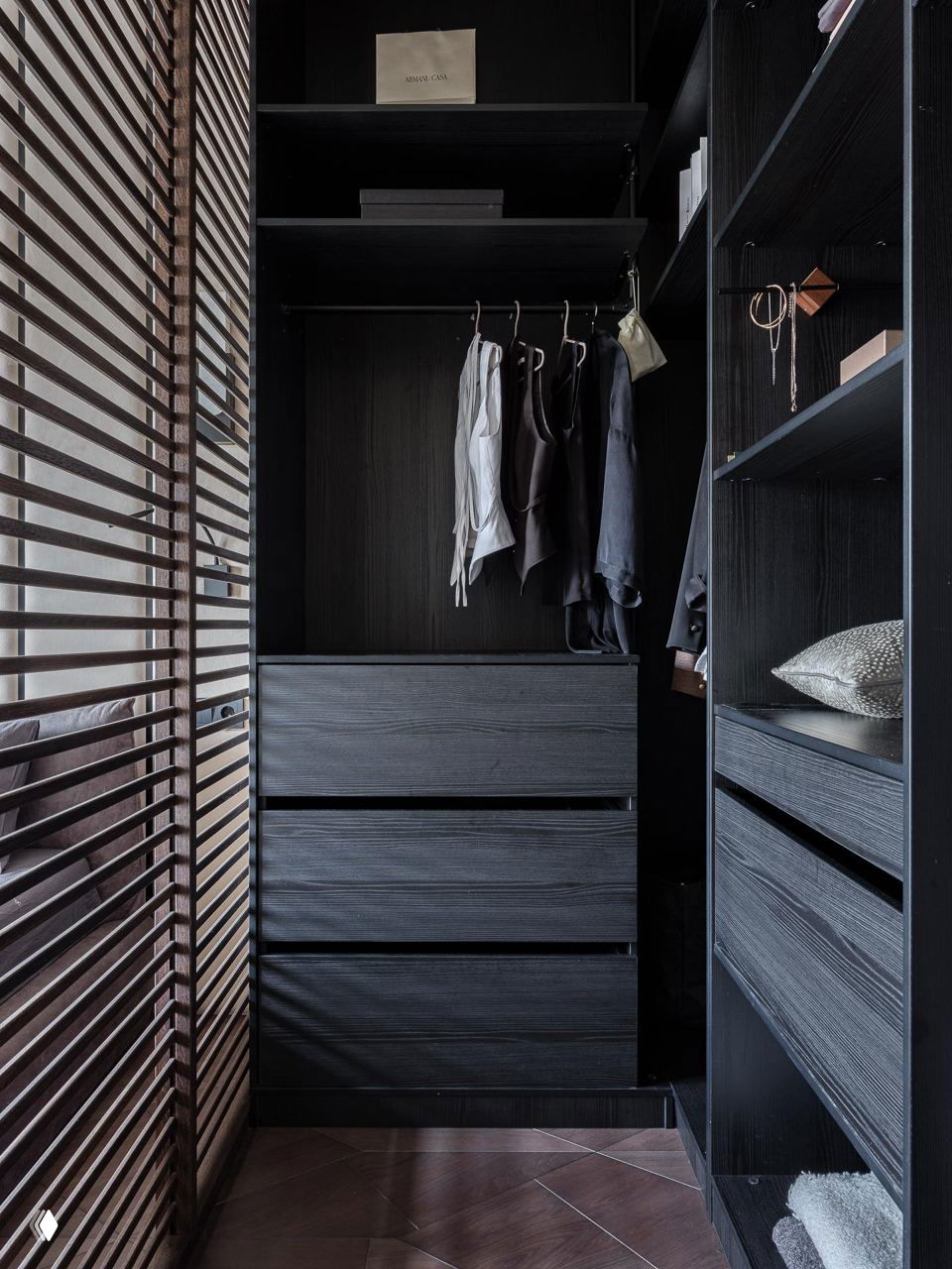 Гардеробная ниша с открытыми полками и ящиками тёмного дерева, аккуратно развешенной одеждой и деревянными реечными ставнями у окна.