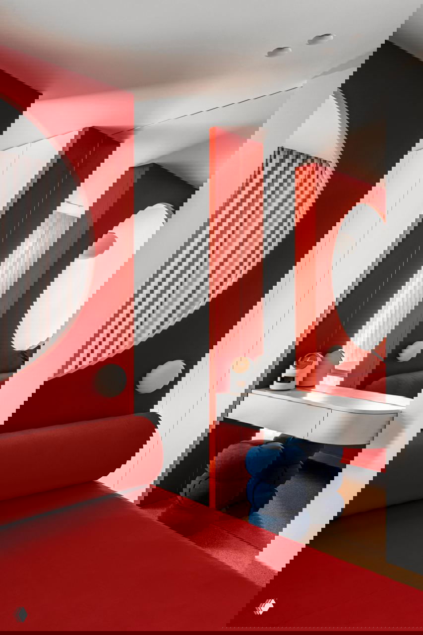 Спальная зона с ярко-красной обивкой, круглым зеркалом и низким столиком, рядом синие пухлые пуфы и встроенные белые шкафы.