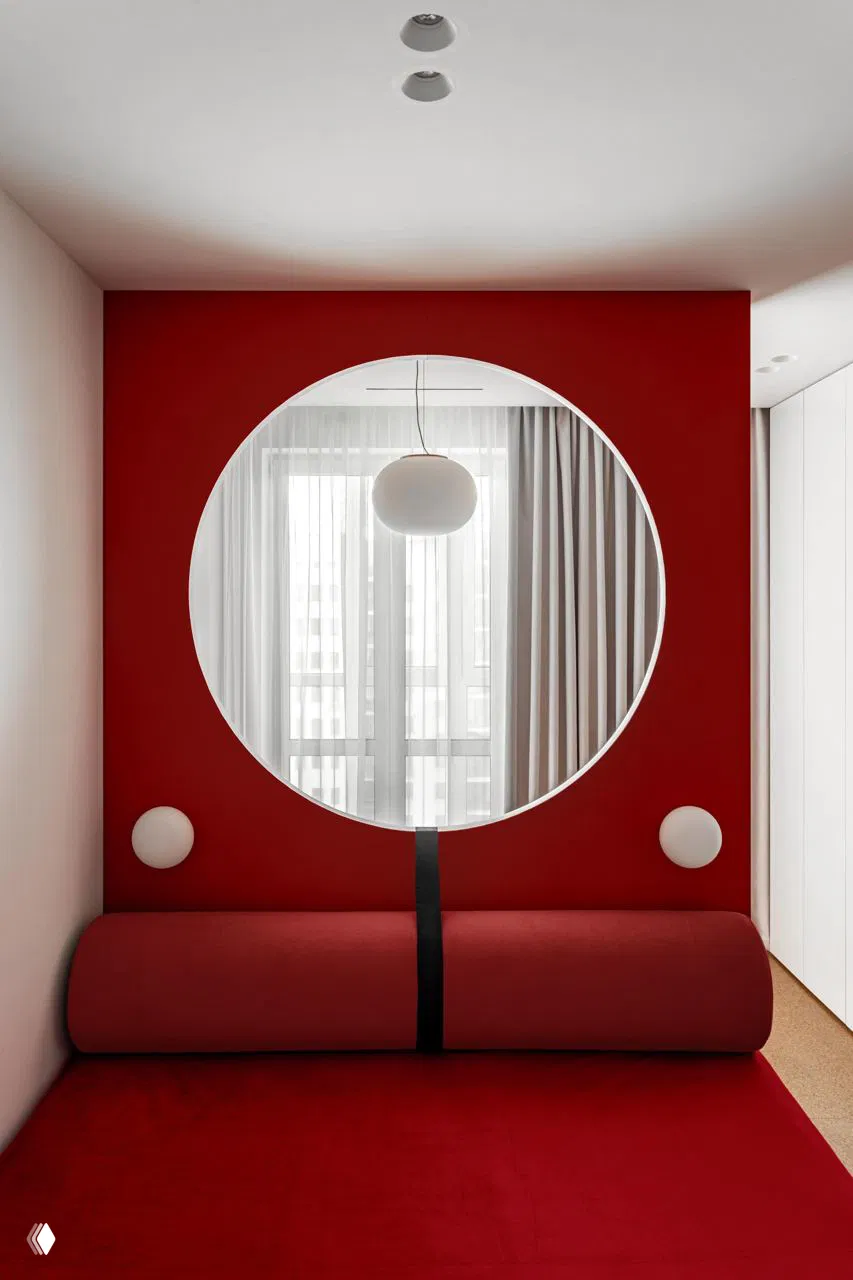 Ниша с большим круглым отверстием в красной стене, через которую видно окно с подвесным светильником; внизу роликовый диван и яркие текстуры.