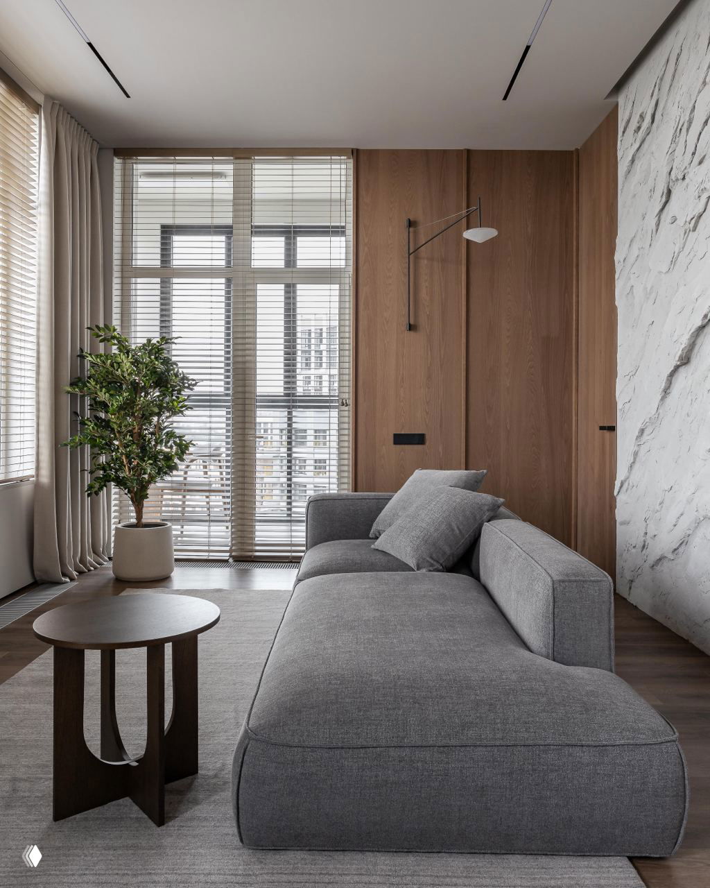 Широкий вид на зону гостиной с серым диваном, деревянные стеновые панели, крупные окна с жалюзи и растением в горшке, атмосфера джапанди.