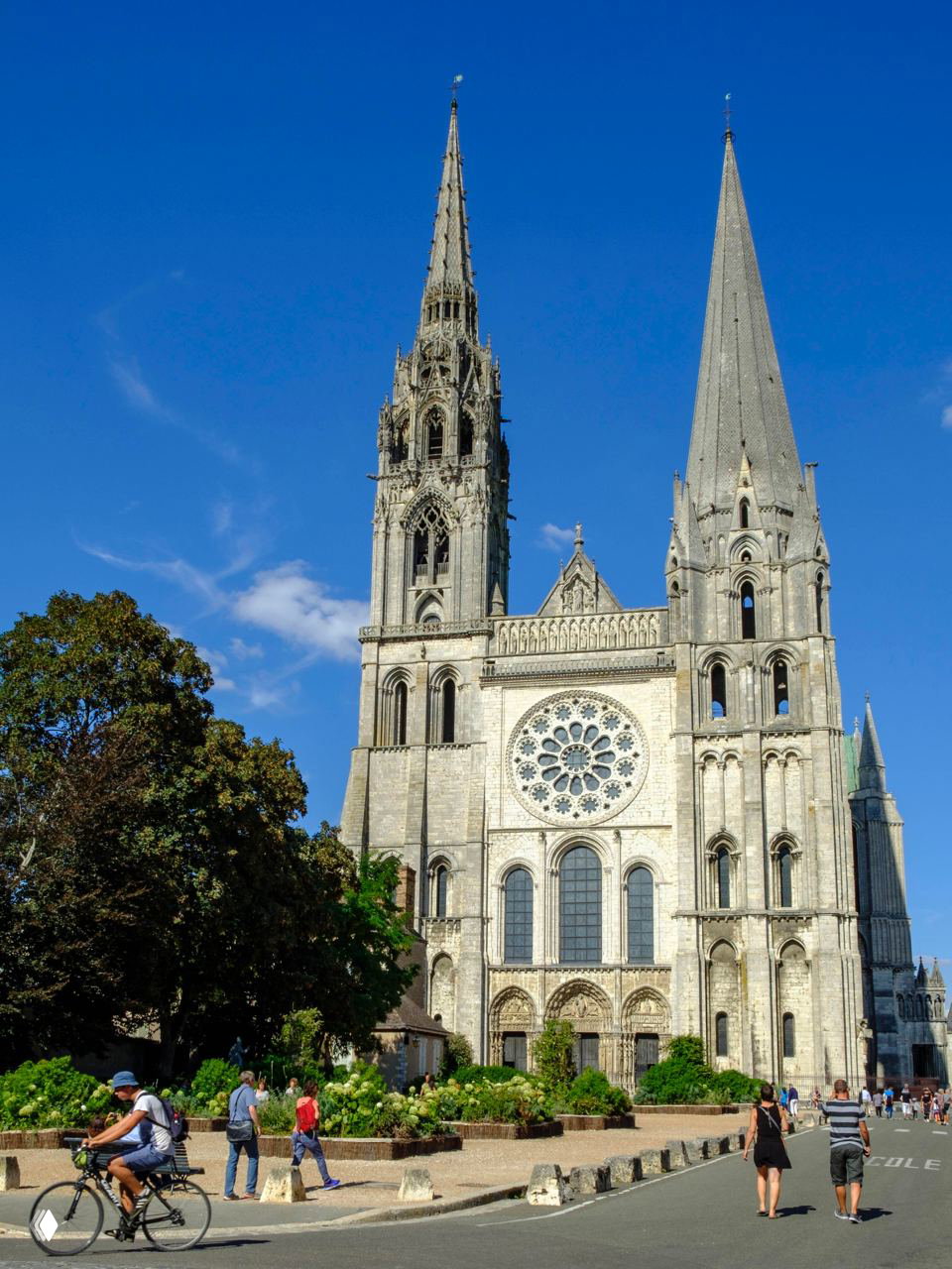 Внешний вид готического собора с двумя асимметричными башнями и розеткой, улица и люди на переднем плане в солнечный день.