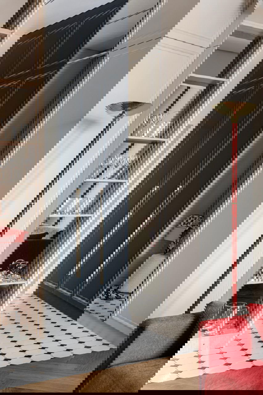 Прихожая и встроенный шкаф тёмно‑синего цвета с зеркальной панелью, на переднем плане красный диван и высокая латунная торшерная лампа, контрастный узор плитки на полу.