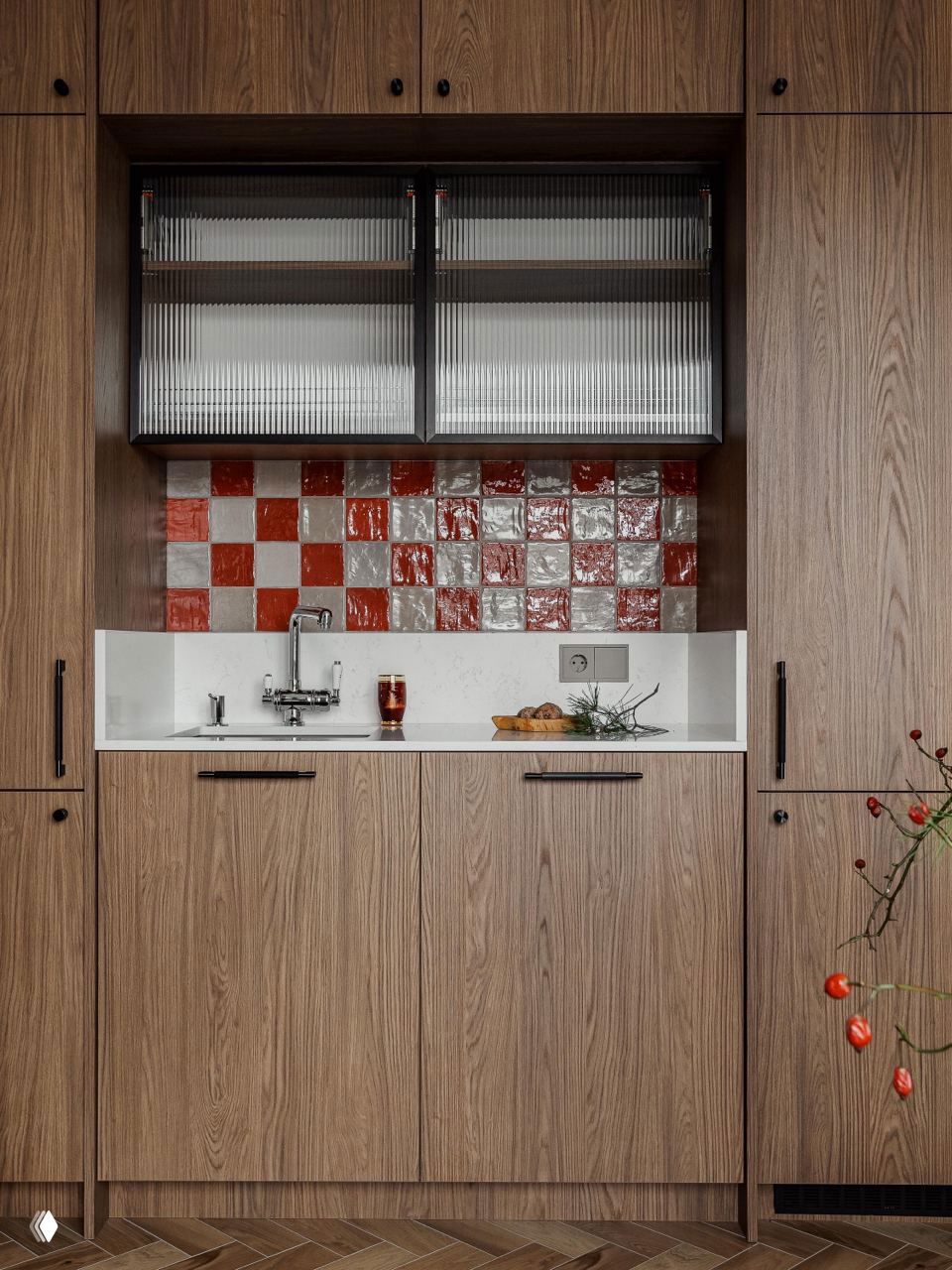 Крупный план кухонного гарнитура из тёплого дерева, белая столешница и глянцевая красно‑бежевая плитка фартука, мойка и небольшой поднос с едой на фоне вертикальных фасадов.
