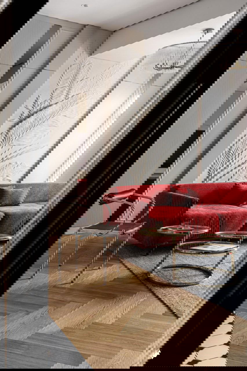 Гостиная с крупным красным диваном на фоне белой рельефной панно в виде больших листьев, журнальные столики с латунными рамами и деревянный ёлочный паркет.