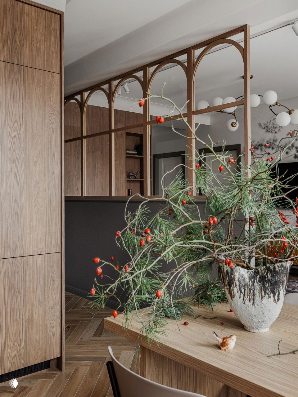 Декоративная деревянная перегородка с арочными нишами между кухней и гостиной; на переднем плане большая ваза с сосновыми ветвями и красными ягодами на деревянном столе.