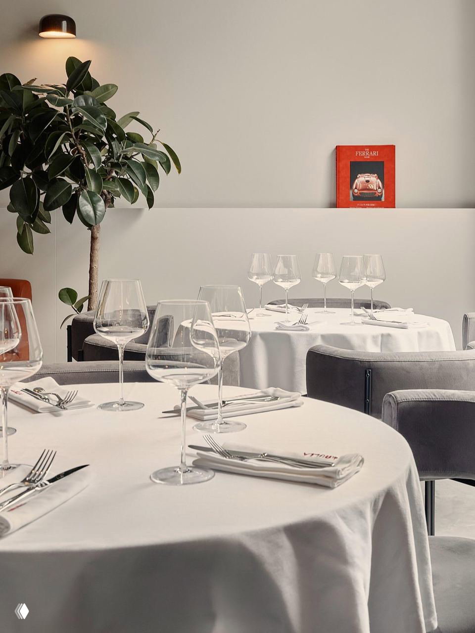 Круглые столы с белыми скатертями, множество бокалов на сервировке и зелёное растение у стены; композиция создаёт ощущение камерности и элегантности.