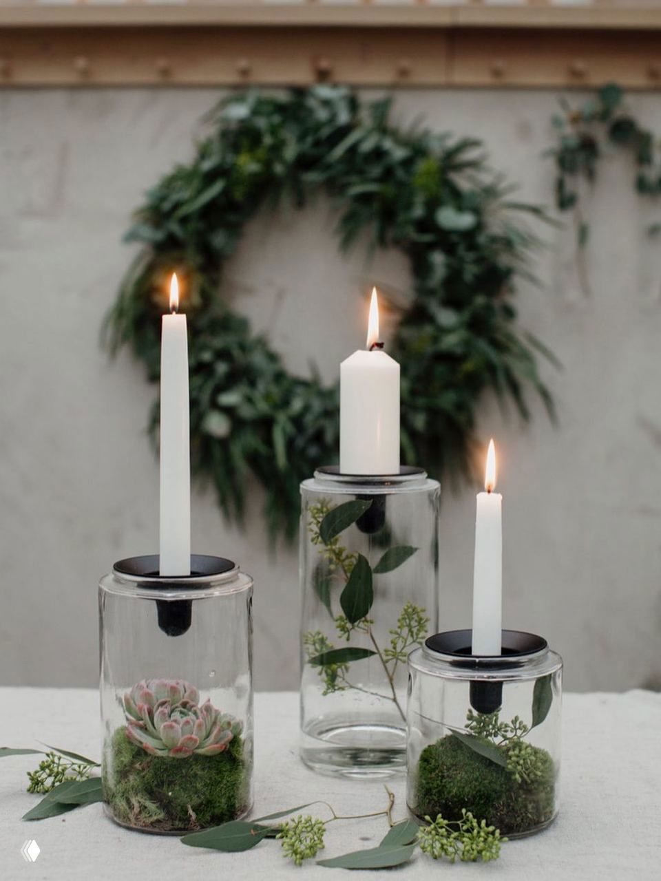 Свечи в стеклянных банках с суккулентами и мхом на переднем плане, на заднем плане размытый венок — минималистичный и естественный настенный декор.