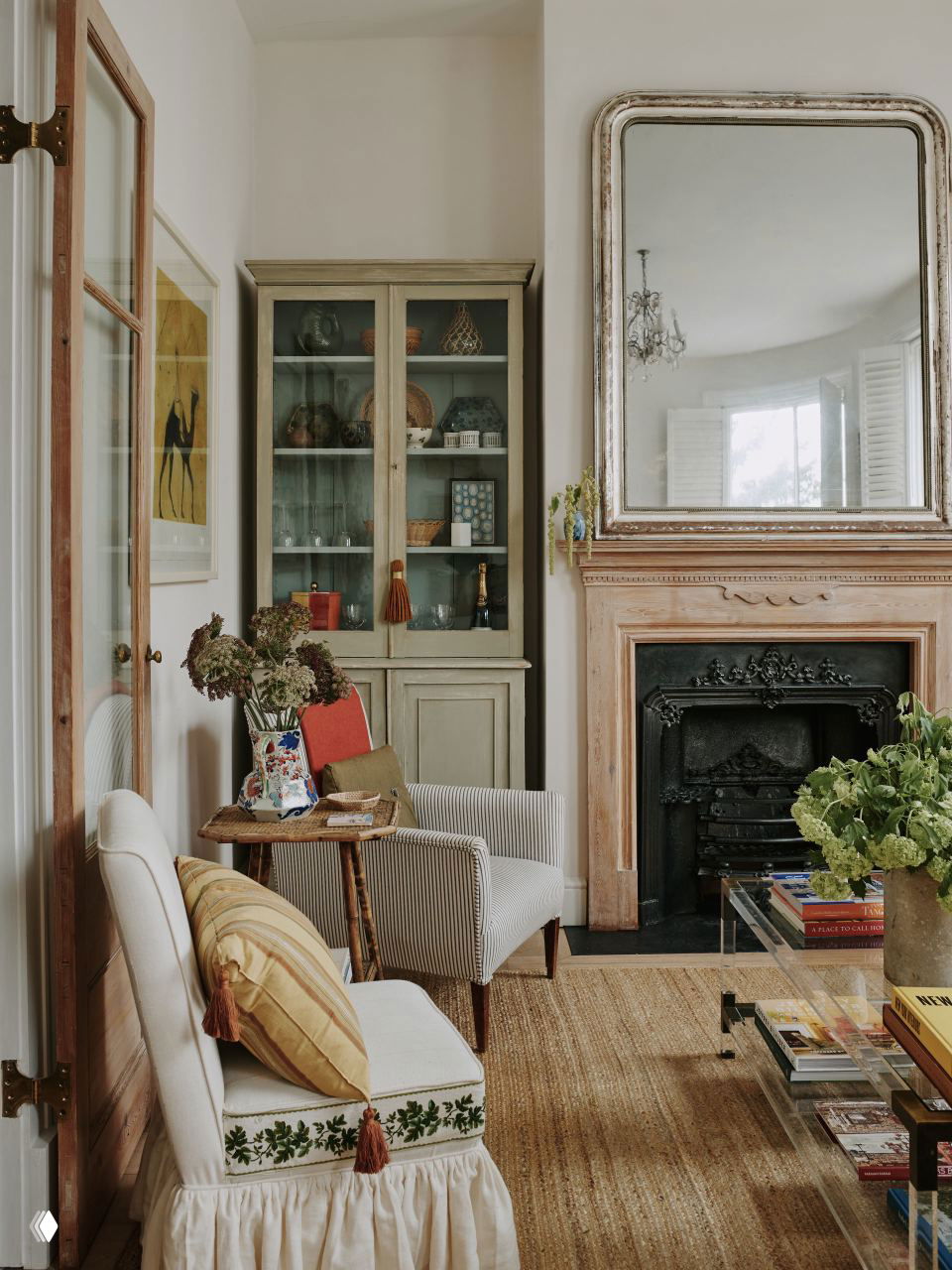 Уютная зона с камином и большим антикварным зеркалом над ним, рядом — витрина с коллекционными предметами и кресло у журнального столика с растением.