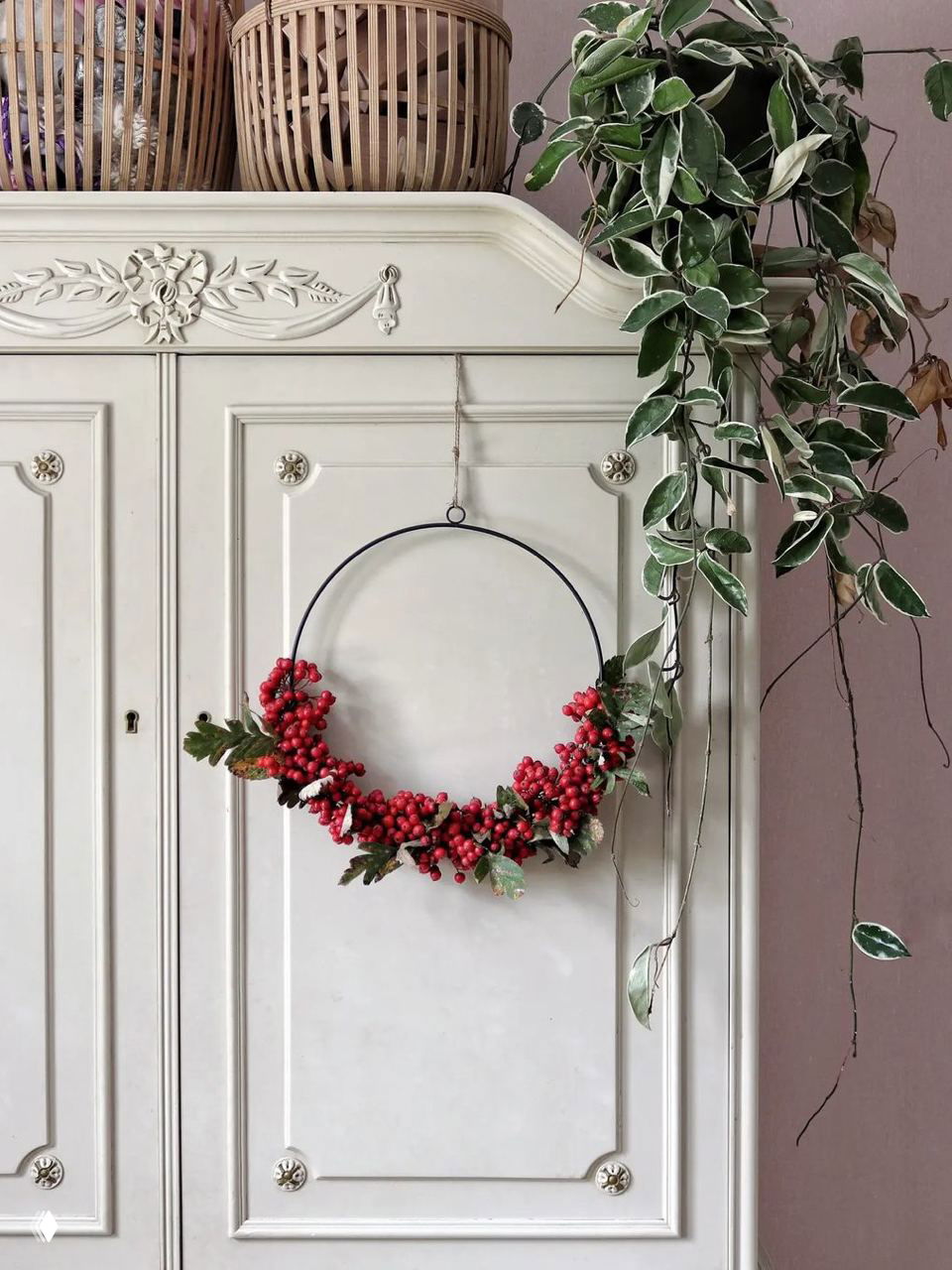 Тонкий металлический обруч с яркими красными ягодами и зеленью на светлом шкафе — лёгкий и воздушный вариант рождественского венка на стене.