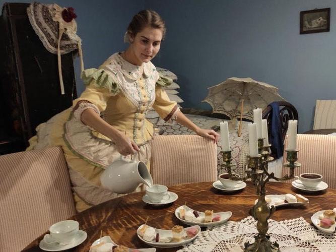 Интерьер музейной экспозиции: женщина в историческом костюме сервирует стол, сцена демонстрирует купеческую кондитерскую традицию Коломны.